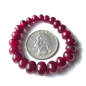May include: A dark red beaded bracelet with a quarter coin next to it for size comparison. The beads are a deep, rich red color and appear to be polished. The quarter shows the profile of George Washington and the year 1998.