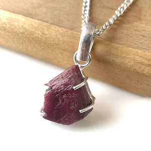 May include: A raw ruby pendant on a silver chain. The ruby is a deep red colour and is held in place by a silver wire setting. The chain is a simple, silver curb chain. The pendant is displayed on a wooden surface.