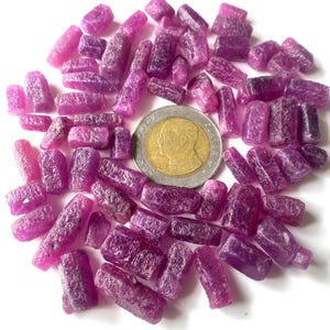 Puede incluir: Una colección de pequeños cristales de rubí rosa en bruto. Los cristales están dispuestos en un círculo alrededor de una moneda de baht tailandesa.
