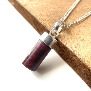 May include: A silver chain necklace with a cylindrical pendant. The pendant is a deep burgundy color with dark spots and silver caps on each end. The necklace is displayed on a wooden surface.