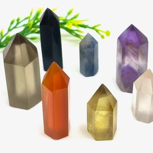 Op de afbeelding: Een verzameling van zeven kleine, puntige kristallen torens in verschillende kleuren, waaronder helder, grijs, zwart, blauw, oranje, geel en paars. De kristallen zijn gerangschikt op een wit oppervlak.