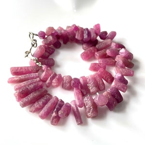 May include: A strand of pink ruby gemstone beads, each bead is a long, thin, rectangular shape with a slightly pointed end. The beads are strung on a silver chain with a clasp.