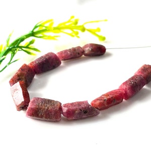 May include: A necklace of rough-cut ruby beads. The beads are a deep pinkish-red colour with natural variations in tone and texture. The beads are strung on a thin, clear wire, and are displayed against a white background with a green and yellow plant.