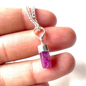 Puede incluir: Un collar de plata con un colgante de rubí en bruto, cilíndrico y magenta. El rubí está engastado en una tapa de plata y cuelga de una delicada cadena de plata. El colgante mide aproximadamente 2 cm de largo.