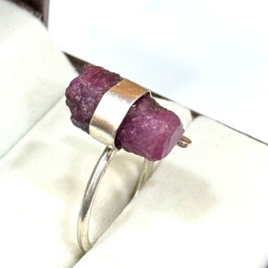 Op de afbeelding: Een zilveren ring met een ruwe robijn edelsteen. De robijn heeft een dieproze kleur en wordt op zijn plaats gehouden door een zilveren band. De band is bevestigd aan een dunne, zilveren ring. De ring staat tegen een witte achtergrond.