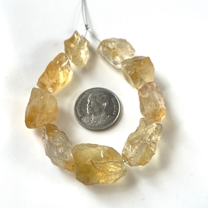 Puede incluir: Un hilo de piedras preciosas de citrino amarillo, crudas y translúcidas, dispuestas en forma circular. Las piedras tienen bordes irregulares y dentados, y tamaños variados. Una moneda de color plateado está colocada en el centro como referencia.