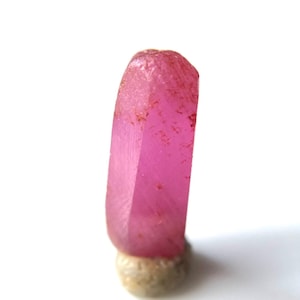 May include: A vibrant pink ruby crystal with a faceted, elongated shape. The crystal is mounted on a small, beige base.