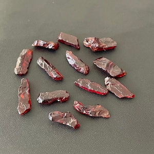May include: A selection of raw, dark red garnet gemstones. The rough-cut stones have irregular shapes and a glossy, reflective surface. The gemstones are scattered on a dark grey surface, highlighting their deep colour and natural texture.