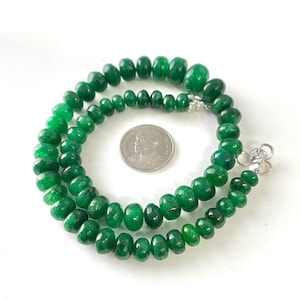 Op de afbeelding: Een ketting van gepolijste, levendige groene kralen. De kralen variëren in grootte en zijn geregen met een zilveren sluiting. De ketting wordt gepresenteerd op een wit oppervlak, met een munt in de buurt voor de schaal.