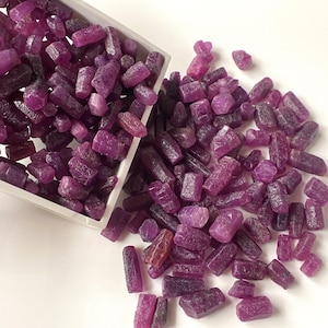 May include: A collection of raw, uncut ruby gemstones in varying shades of deep red and purple. The gemstones are scattered on a white surface, with some spilling from a white rectangular container. The rough-cut stones have a natural, unpolished appearance.