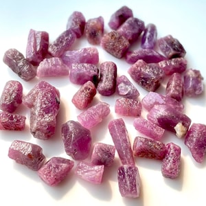 May include: A collection of pink ruby rough stones. The stones are various sizes and shapes, with some being long and thin, while others are more rounded. The stones are all a vibrant pink color.