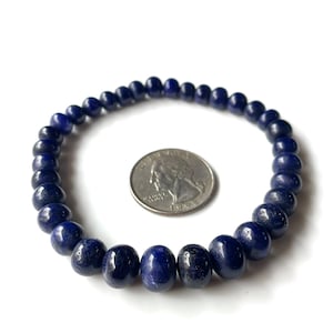 Op de afbeelding: Een donkerblauwe kralenarmband met een munt ernaast ter vergelijking van de grootte. De armband is gemaakt van ronde, gepolijste kralen. De kralen hebben een diepe, verzadigde blauwe kleur. De achtergrond is wit.