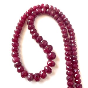 May include: A strand of deep red, faceted, round gemstone beads arranged in a curved shape. The beads have a polished, reflective surface and are strung together, showing a consistent size and color throughout the entire length of the strand.