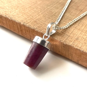 May include: A silver pendant necklace featuring a cylindrical, deep red gemstone. The gemstone is capped with a silver setting and hangs from a delicate silver chain. The necklace is displayed on a wooden surface.