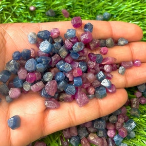 Peut inclure: Une collection de petites pierres précieuses facettées brutes dans des tons de bleu et de rose. Les pierres précieuses sont éparpillées sur une main, certaines se répandant sur une surface verte.