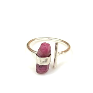 May include: A silver ring with a pink gemstone set in a silver band. The ring has a silver bar extending from the band.