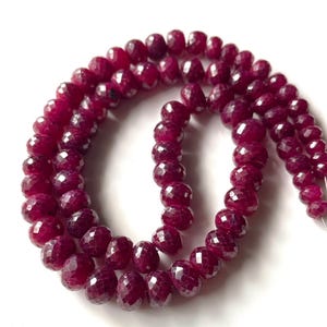 Op de afbeelding: Een streng gefacetteerde, dieprode robijn kralen. De kralen zijn rond en hebben een sprankelend, veelzijdig oppervlak. De kralen zijn aan elkaar geregen, voor een ketting of armband.