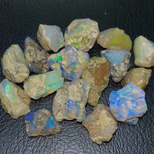 Może przedstawiać: Kolekcja surowych kamieni opalu w różnych odcieniach niebieskiego, zielonego i białego. Kamienie są surowe i mają naturalny, ziemisty wygląd.