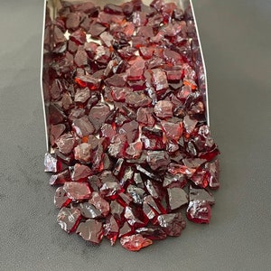 May include: A close-up view of a pile of red garnet gemstones. The garnets are rough and have a variety of shapes and sizes. The garnets are in a metal tray and are spilling out onto a black surface.