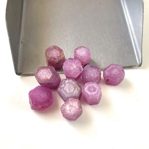 May include: A collection of ten small, hexagonal, raw pink ruby crystals with visible geometric patterns on their top surfaces, resting in front of a metallic scoop. The stones exhibit varying shades of pink and translucent, crystalline textures.