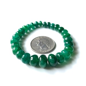 Puede incluir: Un brazalete hecho de cuentas verdes pulidas y vibrantes. Las cuentas están ensartadas, formando una forma circular. Se coloca una moneda de veinticinco centavos estadounidense junto al brazalete para comparar el tamaño. El brazalete está sobre un fondo blanco.