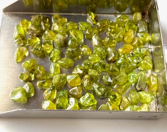 May include: A collection of small, olive-green peridot gemstones displayed in a silver tray. The gemstones vary in shape and size, with a translucent quality. The tray is rectangular with a slightly raised edge, and the background is white.