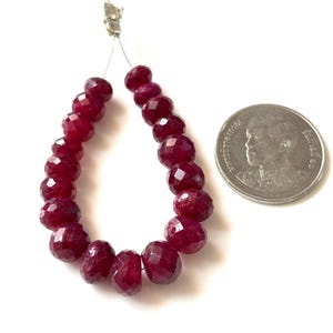 May include: A strand of faceted, deep red ruby beads arranged in a U-shape. The beads are strung on a thin silver-colored wire. A circular silver-colored coin with a portrait of a person is positioned to the right for scale comparison.