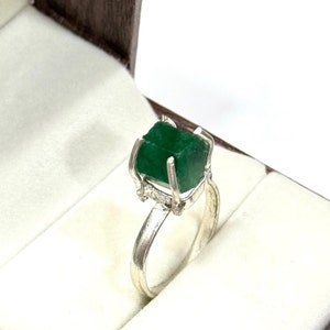 May include: A silver ring with a large, green emerald gemstone set in a claw setting. The ring is in a white velvet ring box.