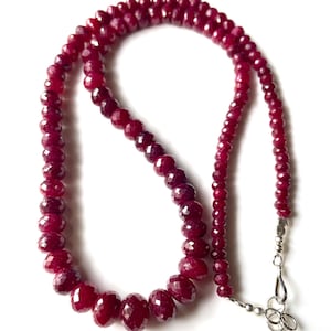 Puede incluir: Un collar compuesto por piedras preciosas facetadas de color rojo intenso. Las cuentas varían en tamaño, con piedras más grandes en la parte delantera y más pequeñas en la trasera. Un cierre plateado asegura el collar. La joya se muestra sobre un fondo blanco.