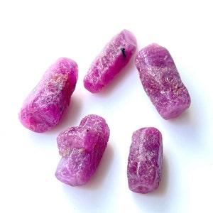 Könnte beinhalten: Fünf rohe Rubin-Edelsteine in einem leuchtenden, rötlich-rosa Farbton. Die Natursteine haben eine leicht strukturierte Oberfläche. Die Edelsteine werden vor einem weißen Hintergrund präsentiert.