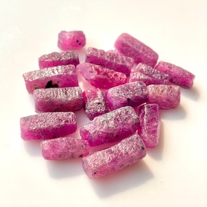 May include: A collection of pink ruby rough stones, some are rectangular in shape, others are more irregular. The stones are all a deep pink colour and have a rough, natural texture.