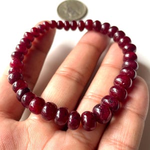May include: A deep red beaded bracelet. The bracelet is made of small, round, polished beads. The beads are a rich, dark red colour. The bracelet is held in a hand against a white background. A coin is visible in the background.