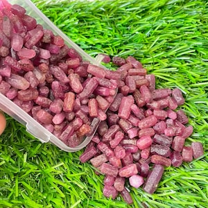 May include: A close-up of a container filled with small, pink ruby crystals. The crystals are scattered on a green, grassy surface.