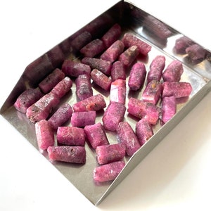 May include: A collection of pink ruby rough stones in a metal tray. The stones are various sizes and shapes, with a rough, natural finish.