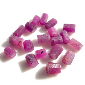 Può includere: Una collezione di gemme di rubino grezze e non tagliate. Le gemme sono di un colore rosso-rosato intenso con una struttura cristallina. Ogni pietra ha una forma e dimensione leggermente diverse.