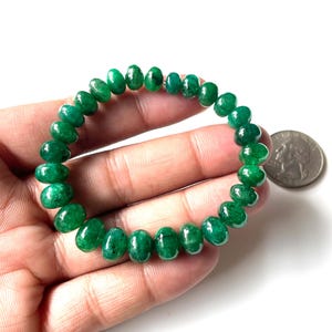 May include: A beaded bracelet featuring vibrant green emerald beads. The bracelet is strung together, forming a circular shape. The beads are polished and have a glossy finish. A US quarter coin is next to the bracelet.