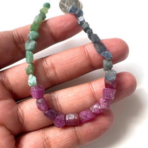 Pode incluir: Um fio de contas de pedras preciosas brutas e não lapidadas dispostas num gradiente de cor. As contas passam do verde claro ao azul profundo e depois a tons vibrantes de rosa e magenta. As pedras têm uma textura rugosa e natural.