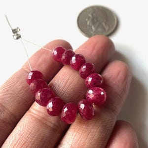 May include: A strand of faceted, deep red ruby beads. The beads are strung on a thin, clear wire. A US quarter is in the background for size comparison. The beads have a sparkling, polished appearance.