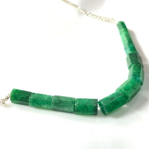 Op de afbeelding: Een zilveren ketting met groene jade kralen. De kralen zijn vierkant en hebben een licht onregelmatige textuur.