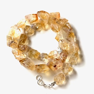 May include: A necklace made of raw, golden-yellow citrine gemstones. The stones are irregularly shaped and strung together with a silver-colored clasp. The necklace is displayed against a white background.