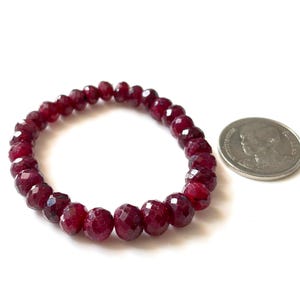 May include: A circular stretch bracelet composed of deep red, faceted, round gemstone beads. The bracelet is positioned next to a silver-colored coin for scale, which measures approximately 1 inch in diameter, providing a clear visual reference.