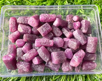 Può includere: Un contenitore di plastica trasparente pieno di pietre preziose di rubino grezze e non tagliate. I rubini sono di un colore rosso-rosato intenso e hanno un aspetto ruvido e strutturato. Il contenitore è su erba verde.