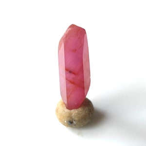 May include: A pink crystal point with a pointed tip and a smooth, polished surface. The crystal is sitting on a small, round, beige stone.