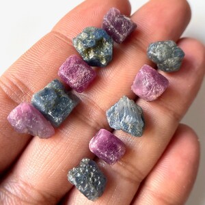 Peut inclure: Une collection de petites pierres précieuses facettées brutes dans des tons de bleu et de rose. Les pierres précieuses sont disposées sur une main, mettant en valeur leur beauté naturelle et leurs formes uniques.