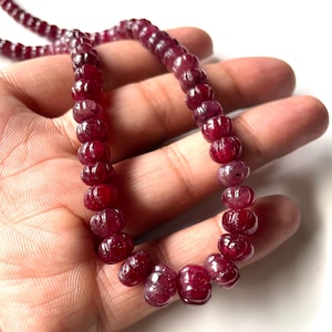 May include: A strand of deep red gemstone beads. The beads are oval-shaped with a carved design, strung together to form a necklace or bracelet. The gemstones have a polished, glossy finish, reflecting light. The overall aesthetic is elegant and natural.