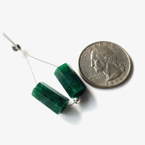 May include: Two faceted, emerald green gemstone beads strung on a silver wire, next to a U.S. quarter for size comparison. The beads are hexagonal in shape and have a polished finish. The quarter shows the profile of George Washington.