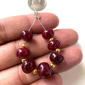 May include: A strand of six polished, deep red, oval-shaped ruby beads alternating with five small, spherical, metallic gold-toned spacer beads, strung on a thin wire, shown next to a U.S. quarter coin for scale reference.