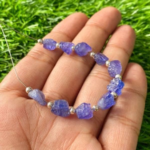 May include: A strand of raw, faceted tanzanite gemstones, each a vibrant shade of violet-blue, interspersed with small, silver-colored beads. The gemstones are strung on a thin, silver-colored wire, creating a necklace or bracelet.