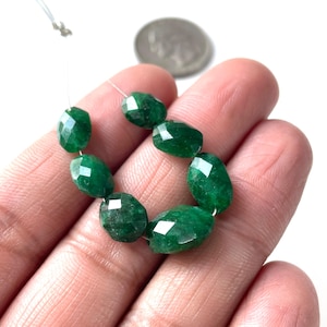 May include: A strand of faceted, oval-shaped emerald green beads. The beads are strung on a thin, clear string. A silver coin is visible in the background.