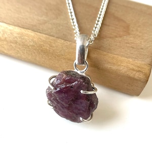 Op de afbeelding: Een zilveren hangerketting met een ruwe, dieprode robijn edelsteen. De robijn wordt vastgehouden in een zilveren kooizetting en hangt aan een fijne zilveren ketting. De edelsteen heeft een ruwe, natuurlijke textuur.
